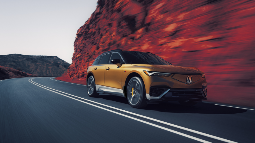 3/4 bird’s eye front view of gold ZDX driving on mountain highway. Part of the image is saturated with colour, so the dirt on the sides of the highway is red.